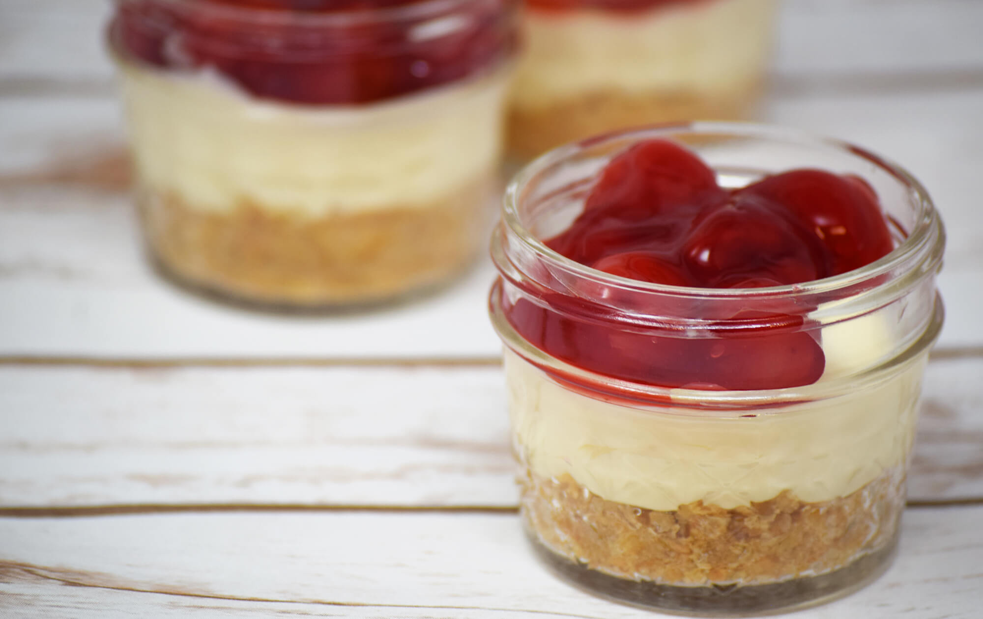 A small mason jar with a prepared cherry cheesecake inside placed on a wood table