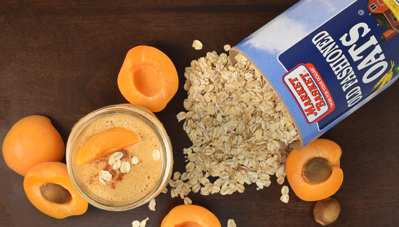 A cinnamon apricot smoothie sits atop a wooden table with fresh apricots slices and Market Basket rolled oats around it