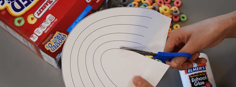 Someone cuts a rainbow pattern from a piece of paper. Elmer's school glue and Market Basket Frosted Fruit O's can be seen on a table in the background.