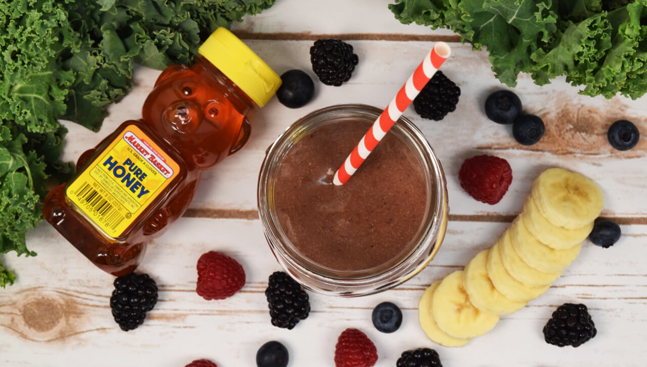 An overhead photo of a smoothie in a mason jar on a wooden backdrop with a bottle of Market Basket Pure Honey and fresh fruit around it