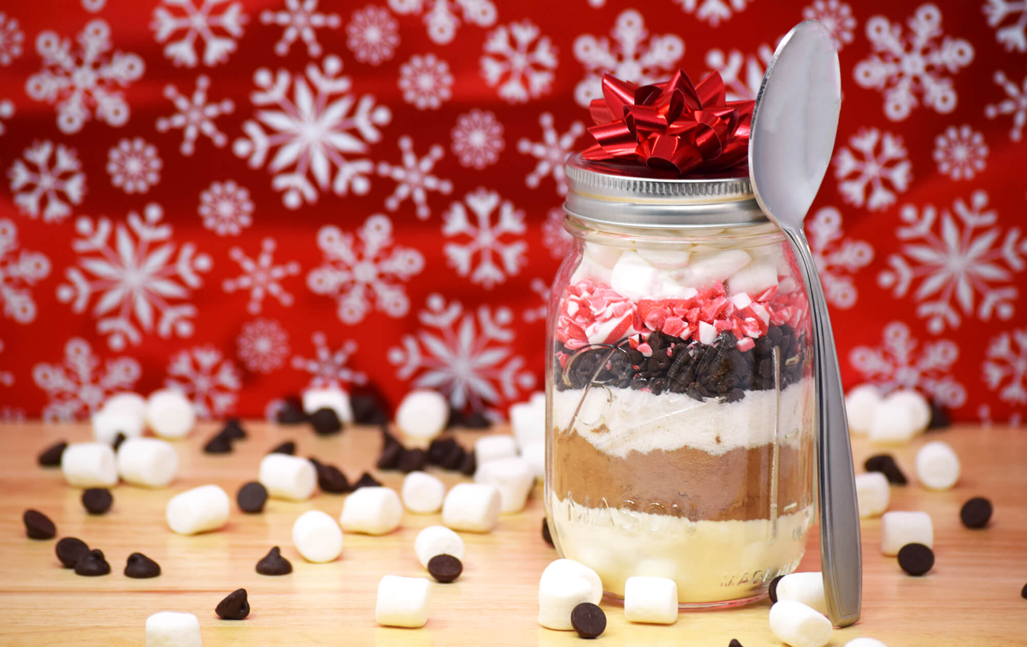 A mason jar with hot cocoa ingredients on a wooden table with marshmallows and chocolate chips surrounding it