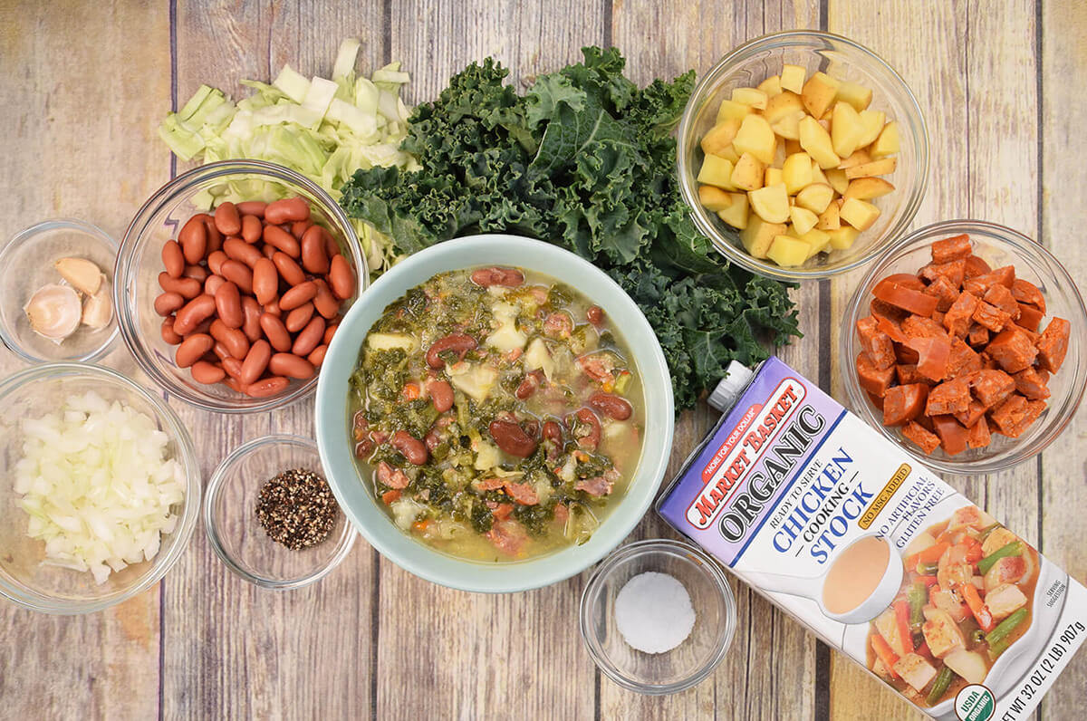 A bowl of kale and chourico soup sits atop a wooden background surrounded by small glass bowls filled with fresh ingredients such as kidney beans, onions, potatoes, and chorizo as well as a container of Market Basket chicken stock