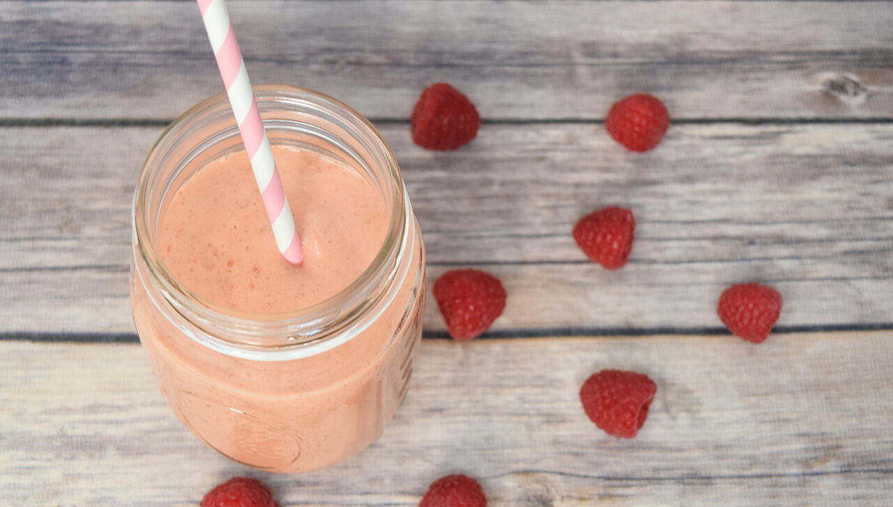 A mason jar filled with a pink colored smoothie sits on a wooden background with raspberries scattered around it