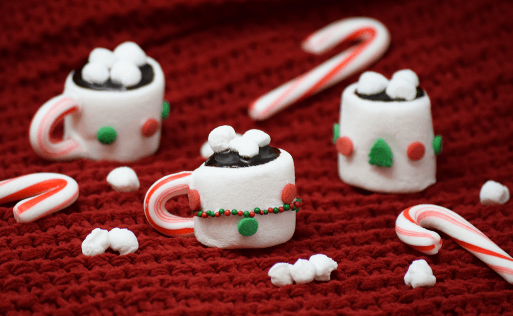 Miniature hot cocoa mugs made from marshmallows, sprinkles, candy canes, and chocolate placed on a red blanket