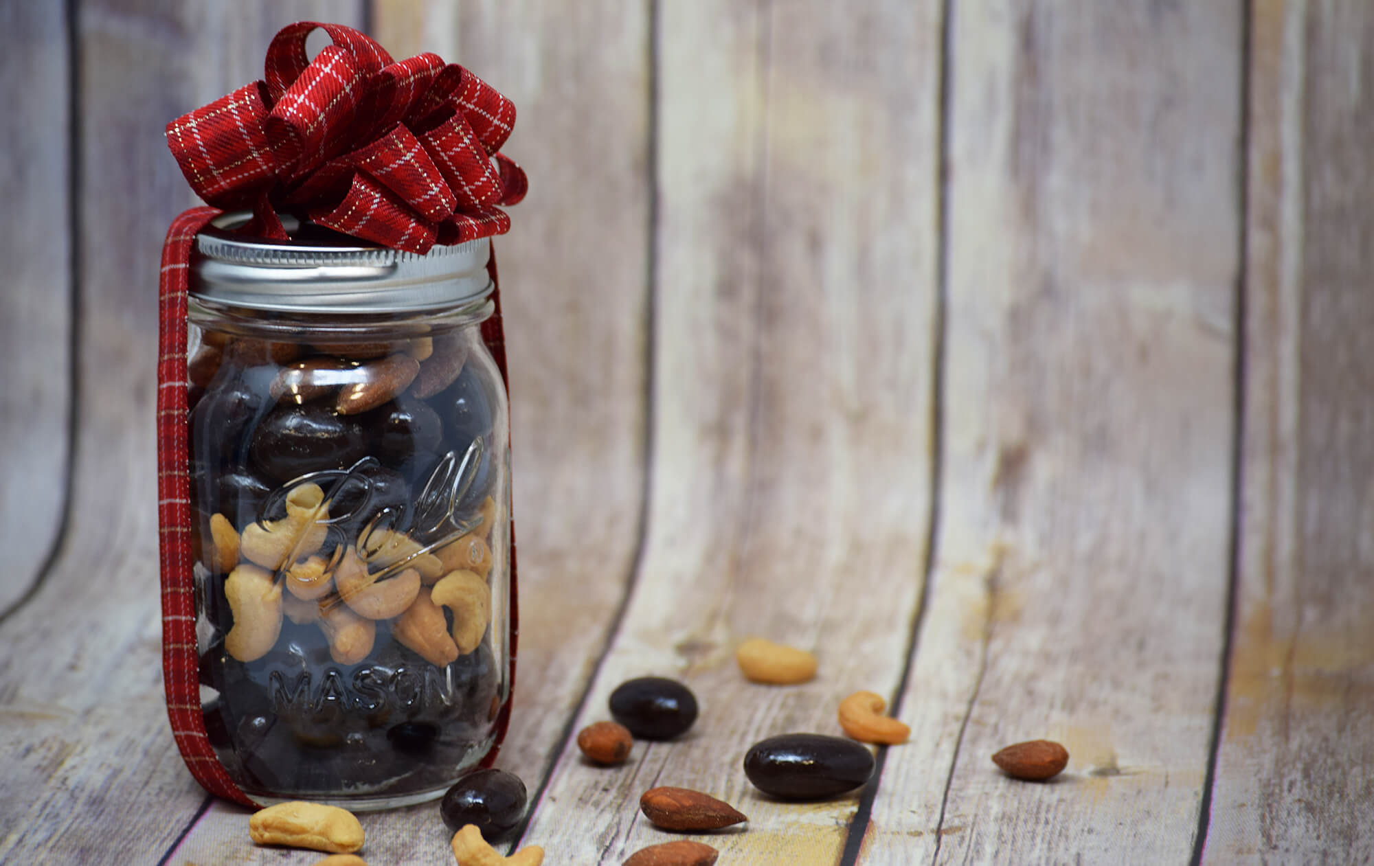 A mason jar filled with mixed nuts and chocolate tied with a bow on a wooden backdrop