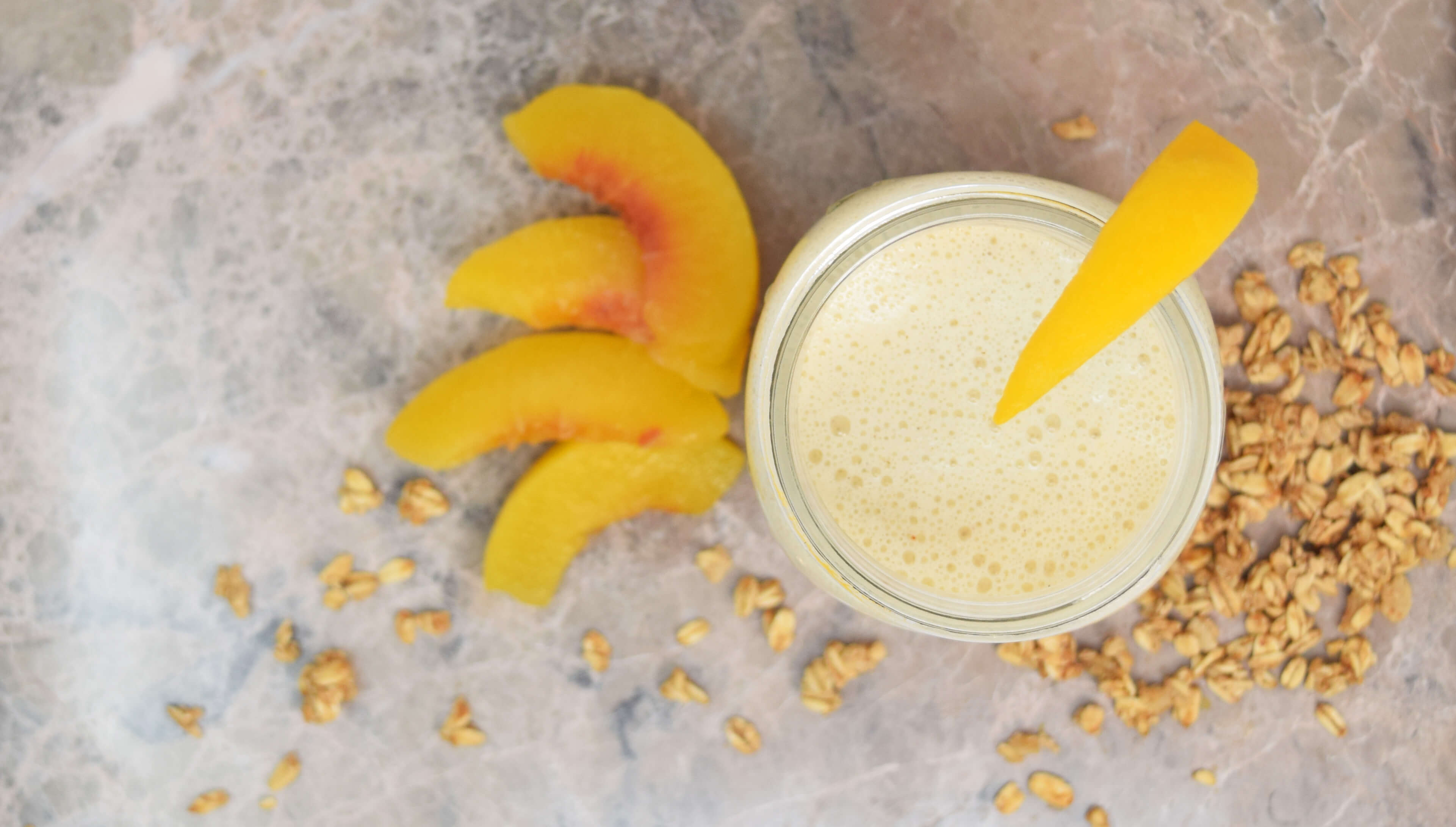 An overhead shot of a peach cobbler smoothie inside a mason jar on a marble counter. Around the mason jar are freshly sliced peaches and granola.