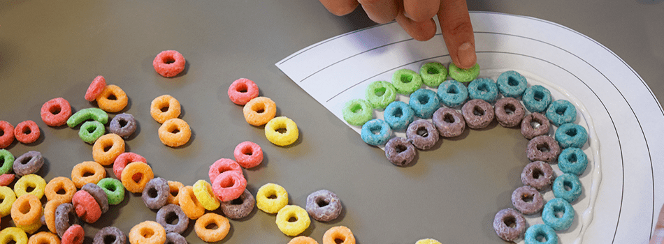 A hand placing a Market Basket Frosted Fruit O's cereal piece onto the rainbow template