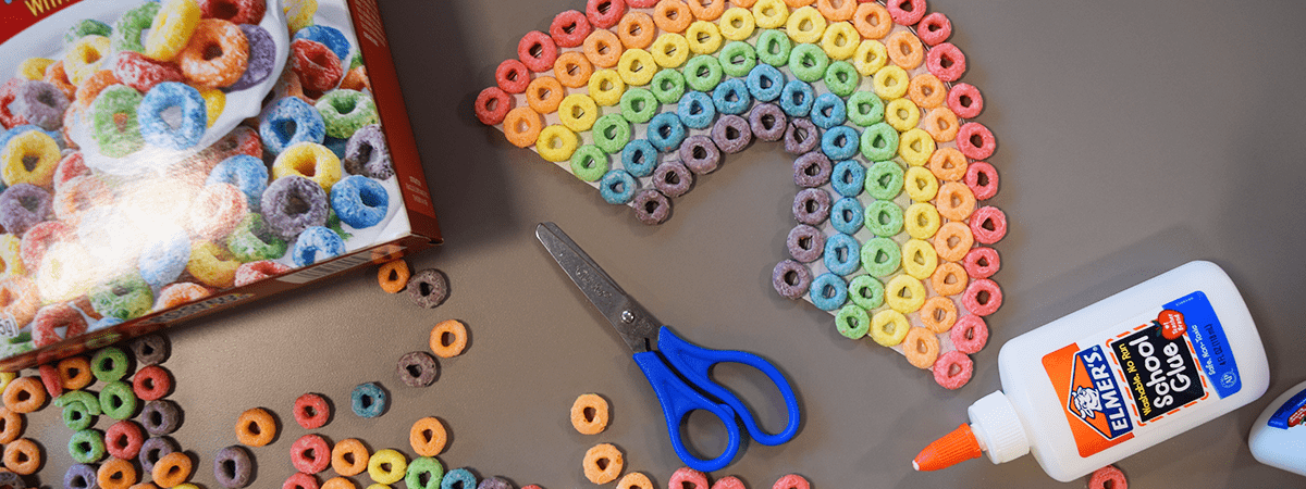 A rainbow made with Market Basket Frosted Fruit O's cereal sits atop a table surrounded by Elmer's school glue, a cereal box, safety scissors, and loose cereal pieces
