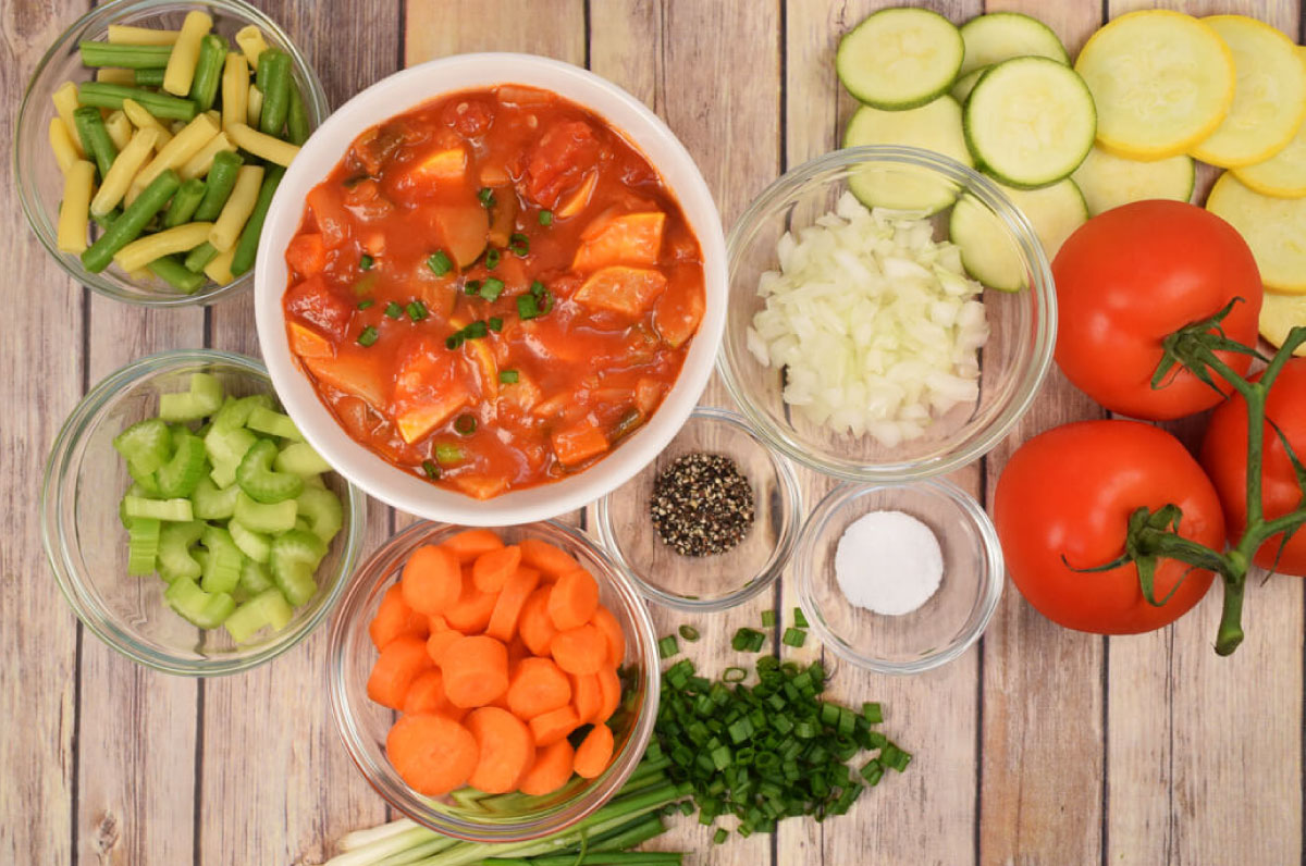 A bowl of tomato vegetable soup sits on a wooden counter with fresh vegetables and chopped or sliced ingredients in small glass bowls around it