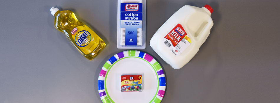 An overhead shot of the supplies needed to make a milk kaleidoscope: dish soap, cotton swabs, whole milk, a plate, and food coloring