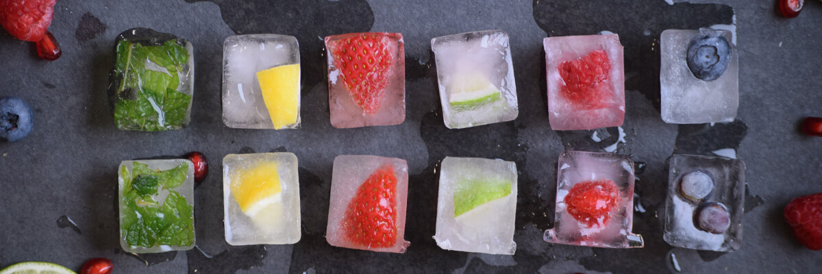 Fresh fruit, vegetables, and herbs frozen in ice cubes that are set up symmetrically on a slate background